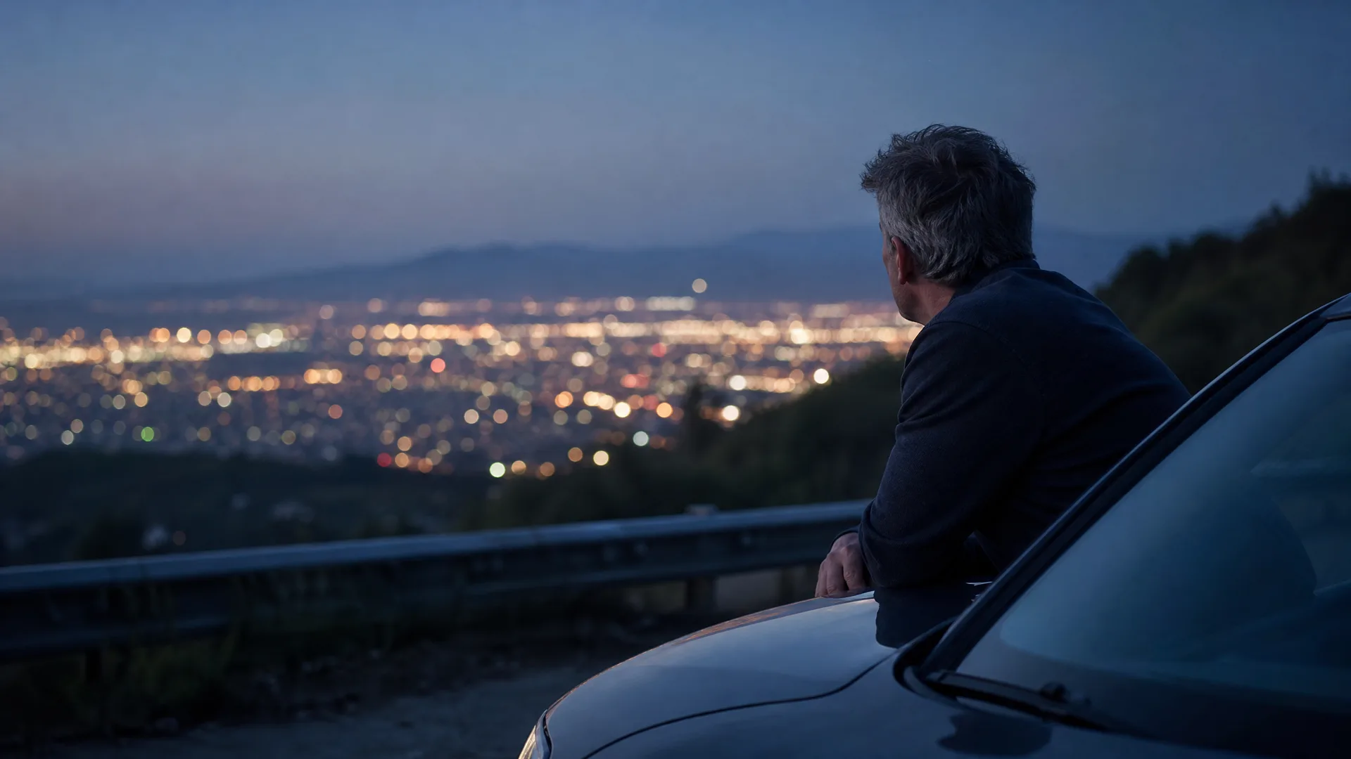 A man in a dark jacket leans on the hood of a car at a roadside overlook at dusk, facing a valley filled with city lights below distant hills.