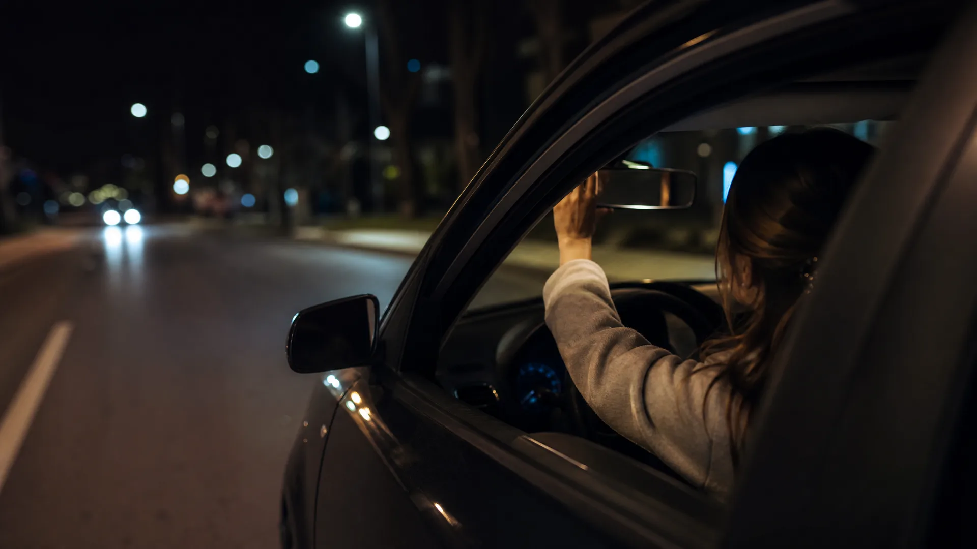 A woman seen through an open car window at night, reaching up to adjust the rear-view mirror while seated at the steering wheel, with a dark street and distant headlights and streetlights to the left.