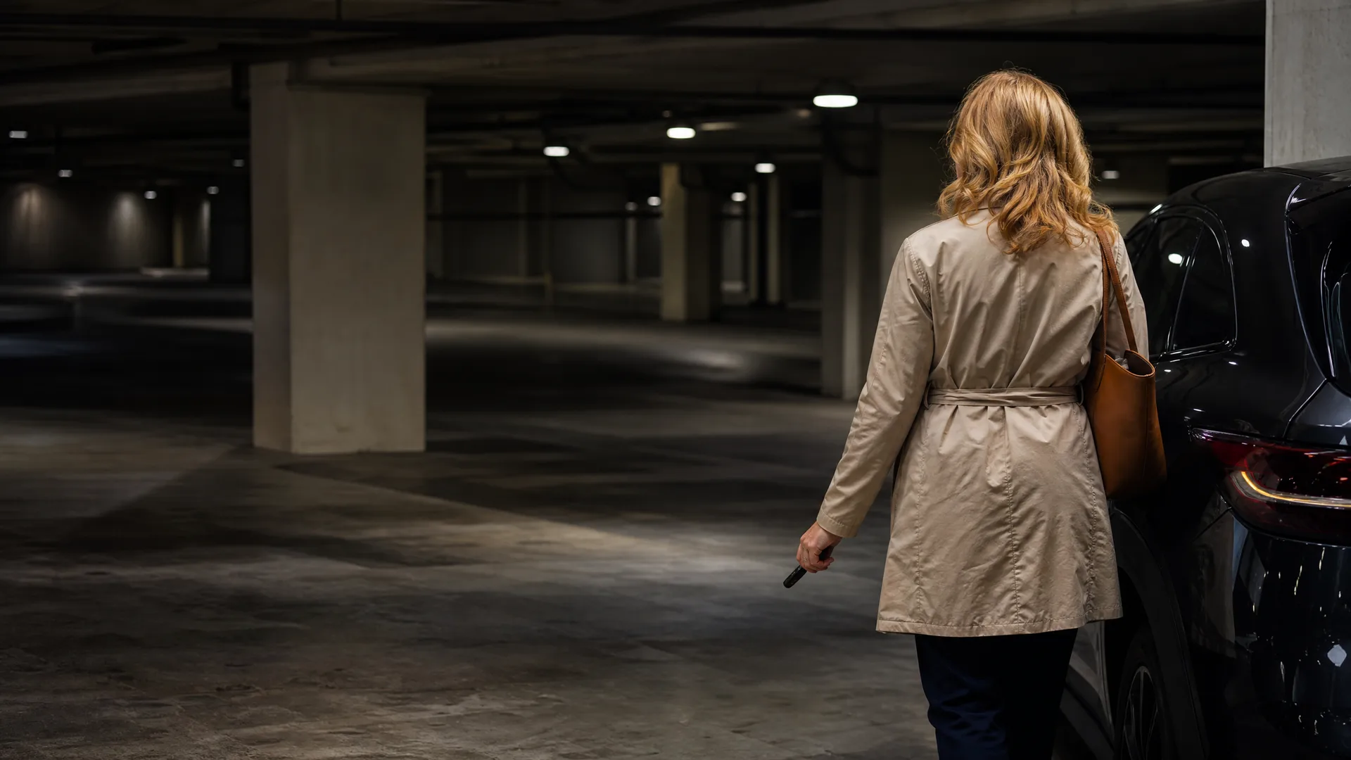 A woman in a beige trench coat with a tan shoulder bag walks toward a dark parked car in a dimly lit concrete parking garage, seen from behind.