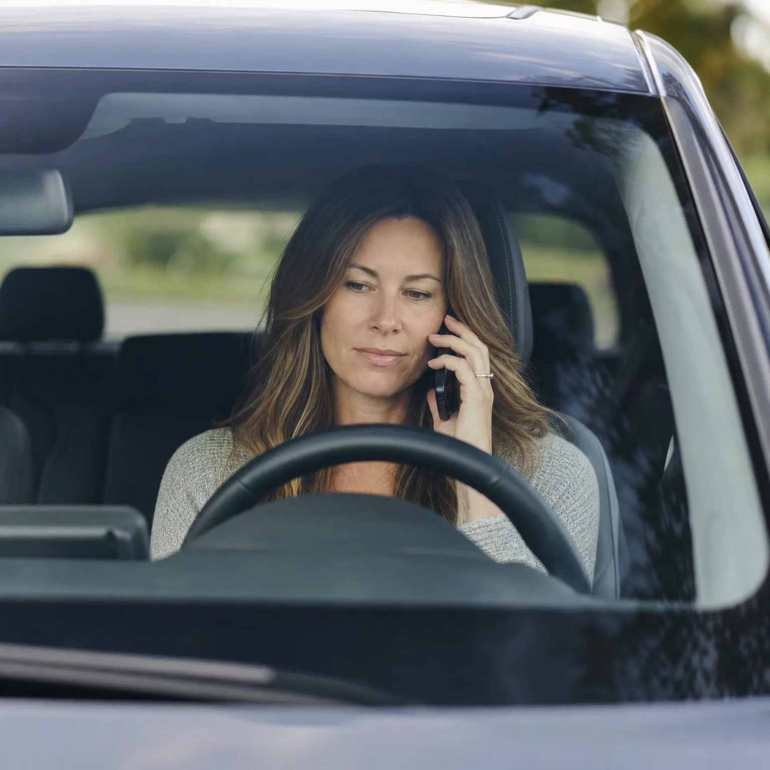 A woman with long brown hair sits in the driver's seat of a car holding a phone to her ear, seen through the windshield above the steering wheel.