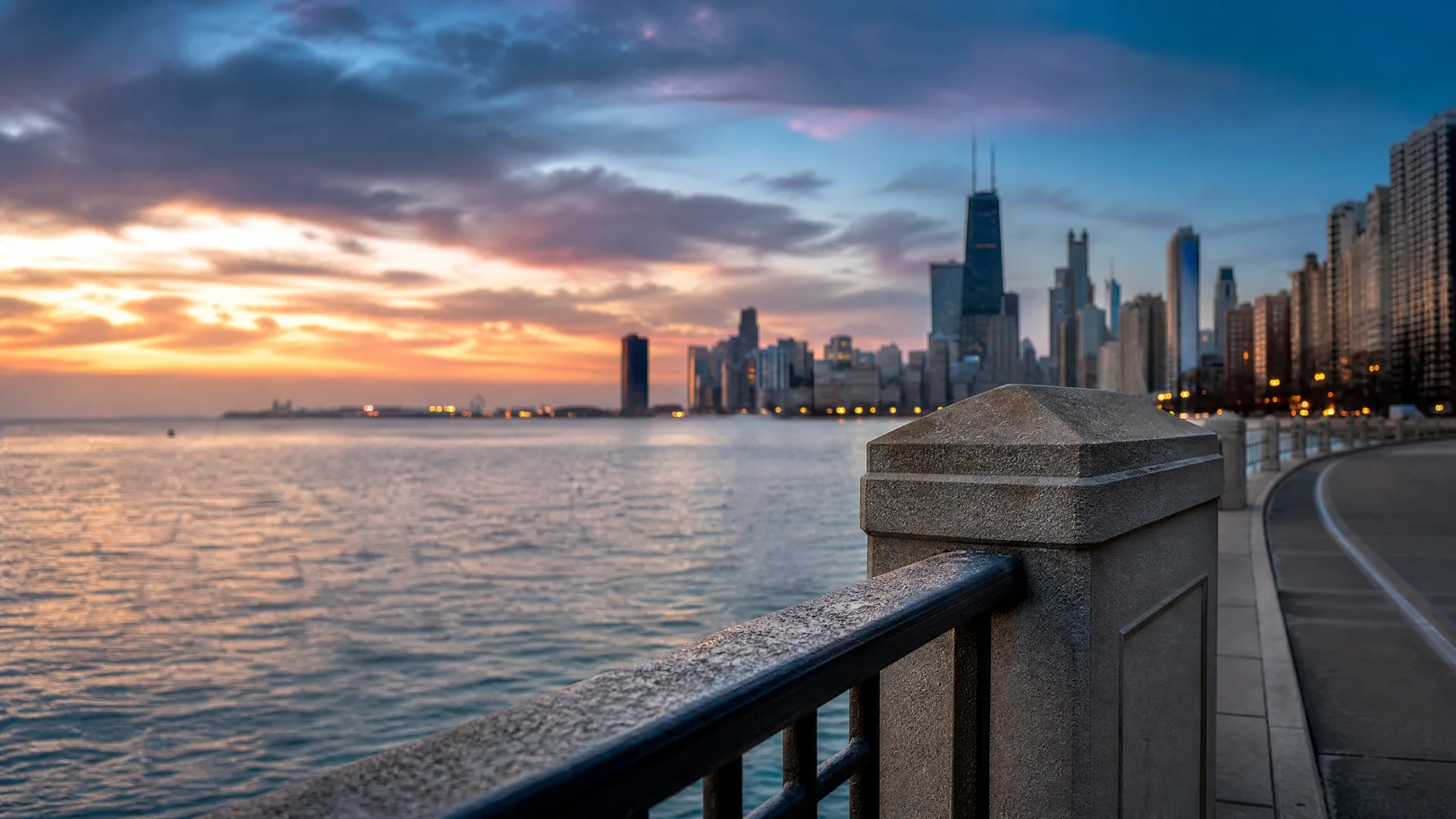 A stone and metal railing with a square post in the foreground along a curved waterfront walkway, with a large body of water and a city skyline under a cloudy sunset sky.