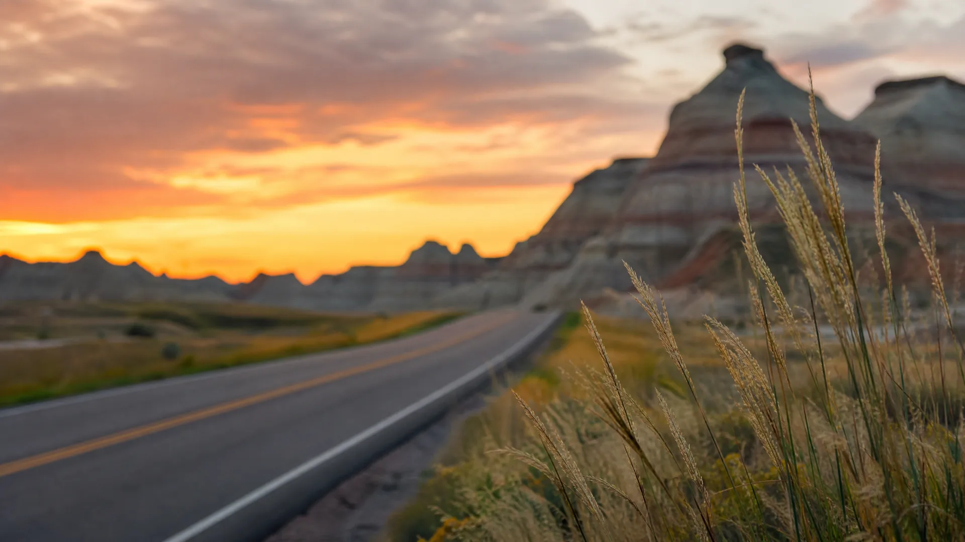 A two-lane road with a yellow center line curves through open terrain at sunset, with tall golden grasses in sharp focus in the right foreground and blurred eroded rock formations and an orange sky behind.